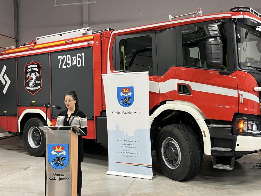 Kobieta przy mównicy na tle samochodu.