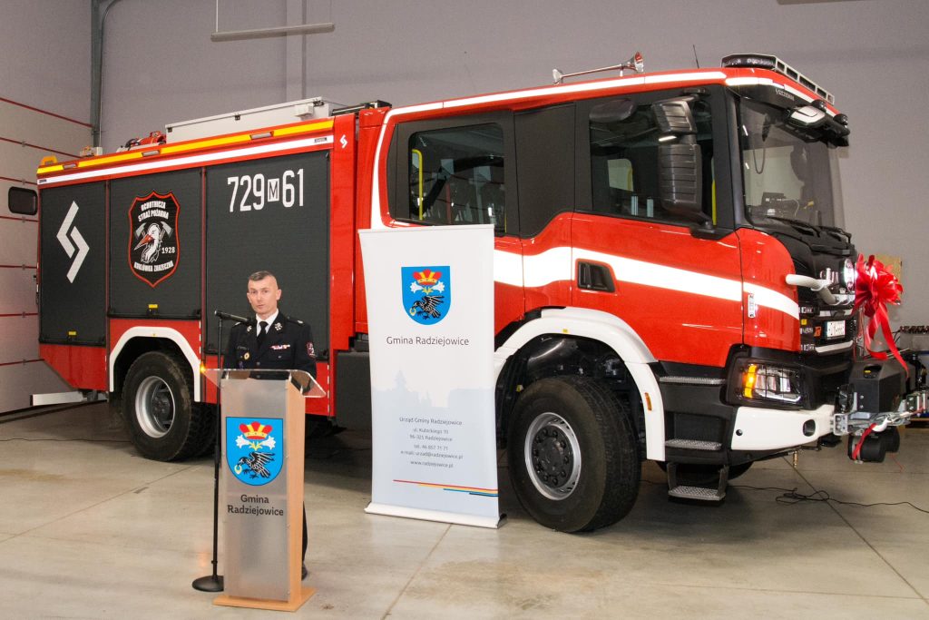 Strażak przy mównicy na tle samochodu.