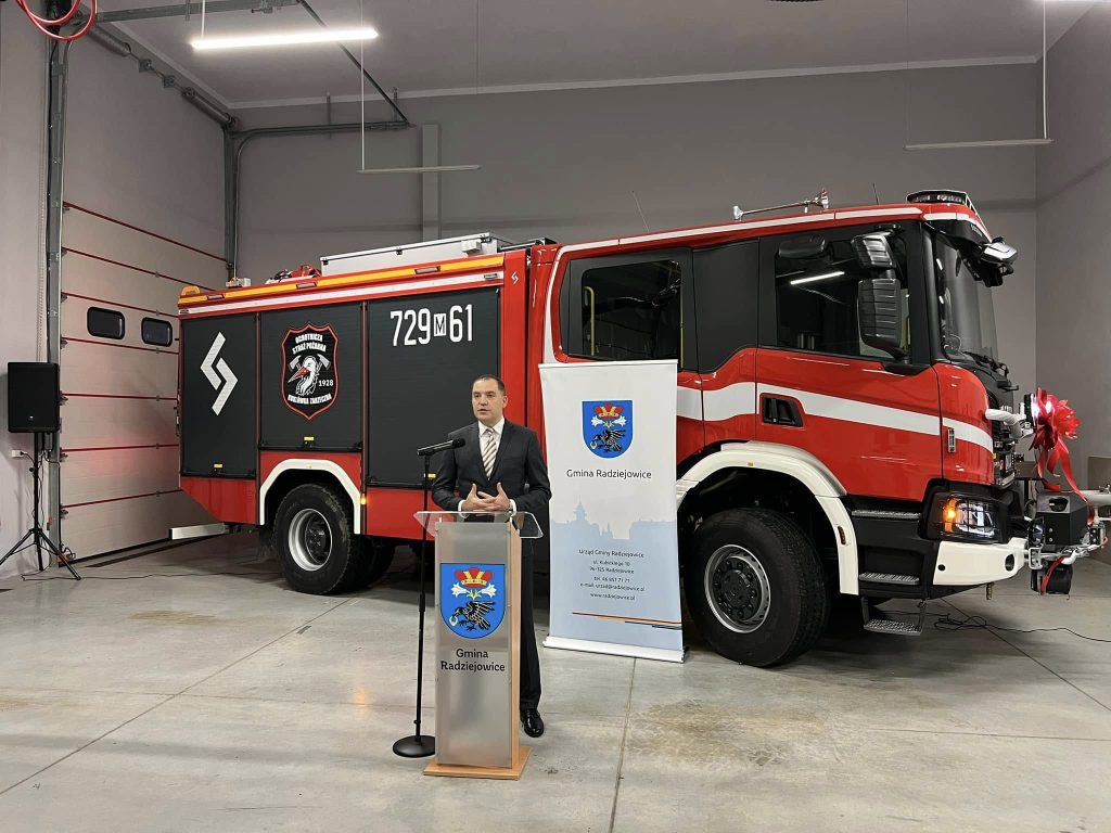 Mężczyzna przy mównicy na tle samochodu.