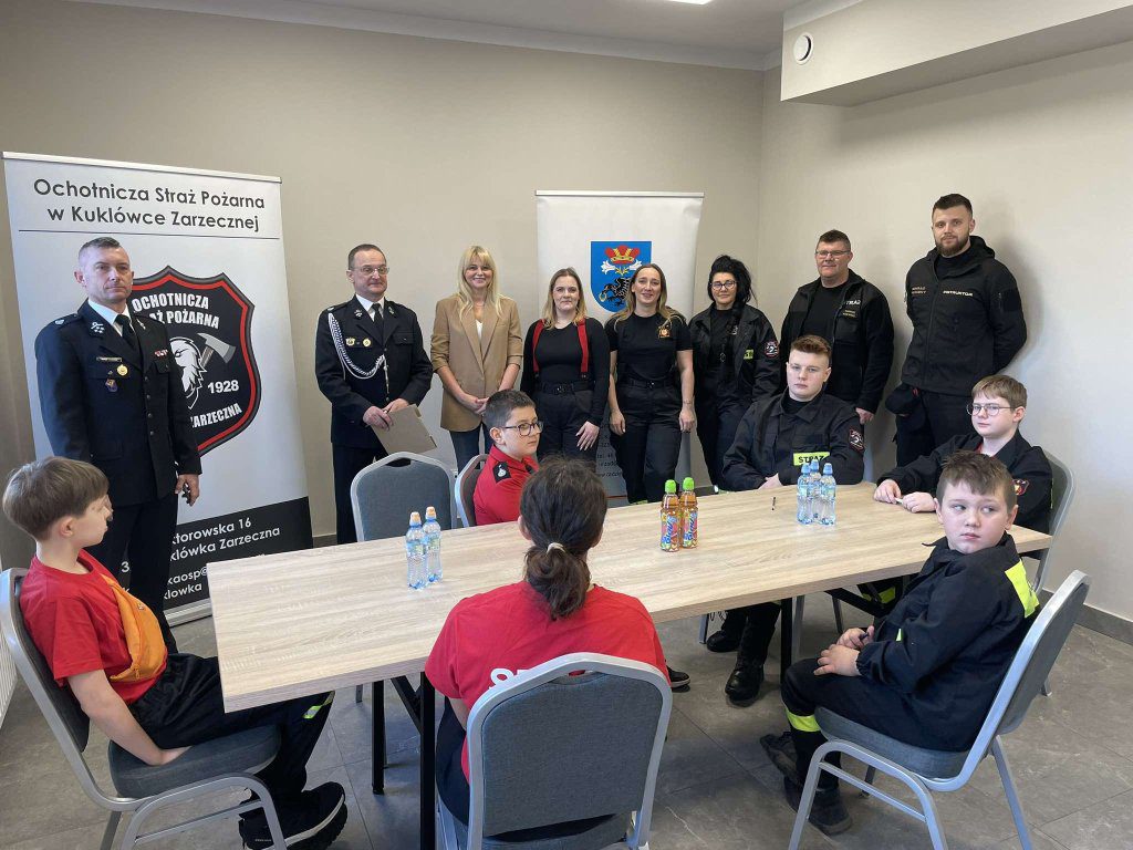 Młodzież siedzi przy stole. Grupa dorosłych osób stoi z tyłu.