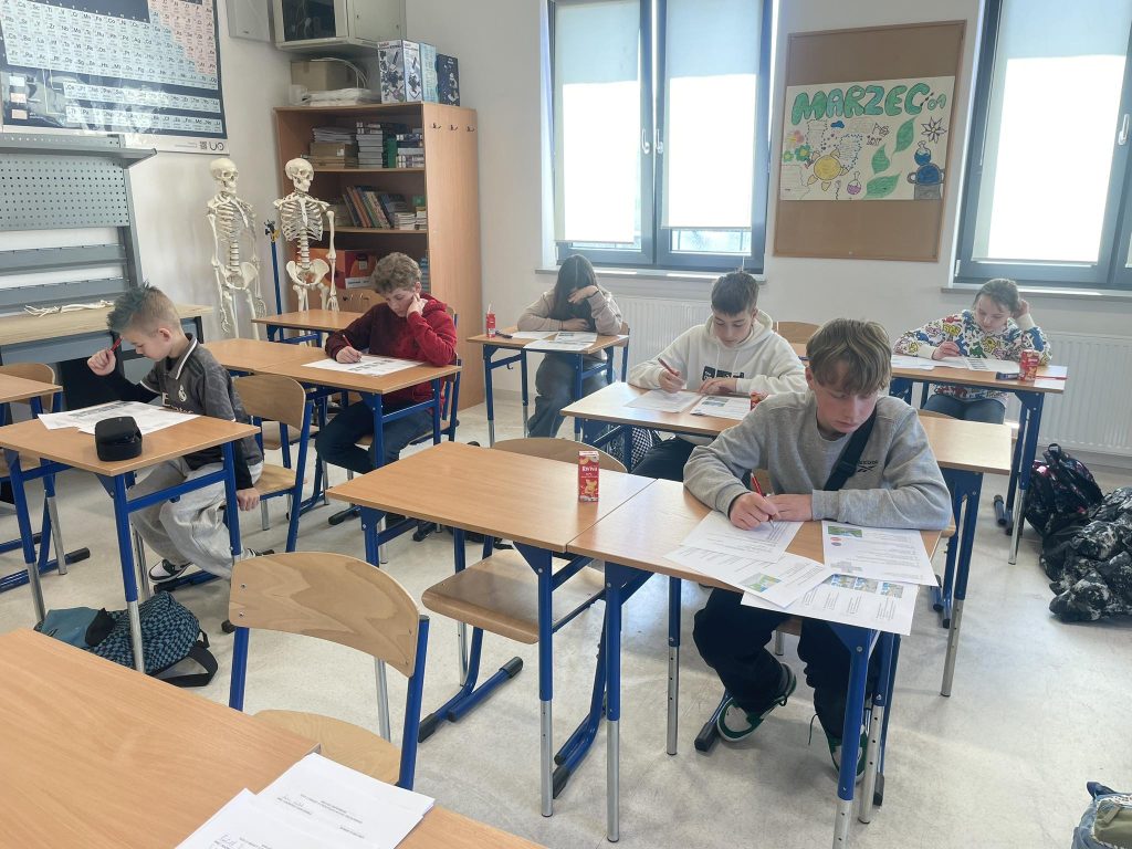 Dzieci siedzą w ławkach i piszą