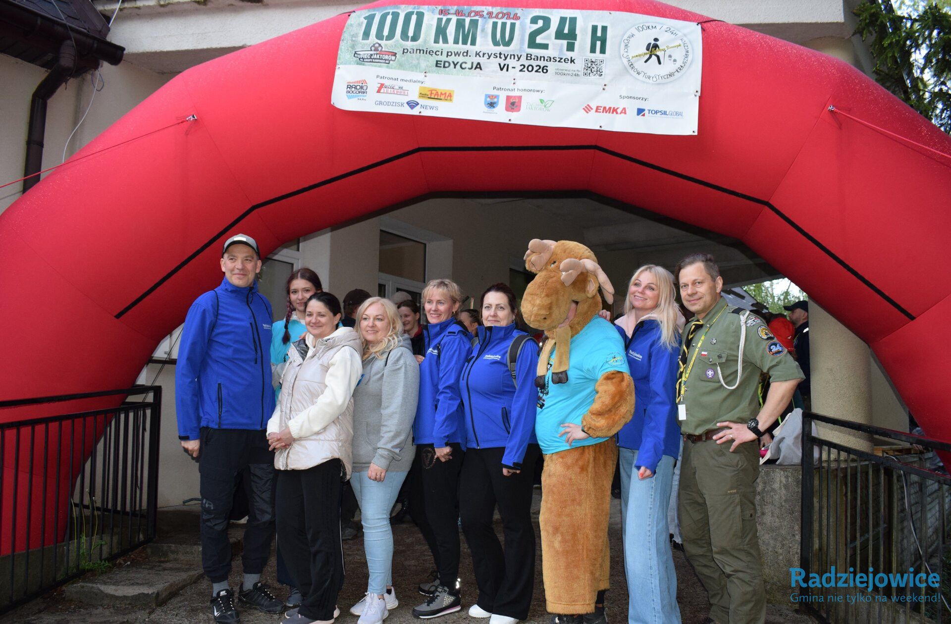 Grupa uczestników, w tym przedstawiciele samorządu Gminy Radziejowice oraz organizatorzy Marszu 100 km w 24 h pamięci pwd. Krystyny Banaszek, pozuje do wspólnego zdjęcia pod czerwonym łukiem startowym z banerem VI edycji wydarzenia. Wśród osób znajduje się uczestnik w kostiumie łosia oraz osoba w mundurze harcerskim.