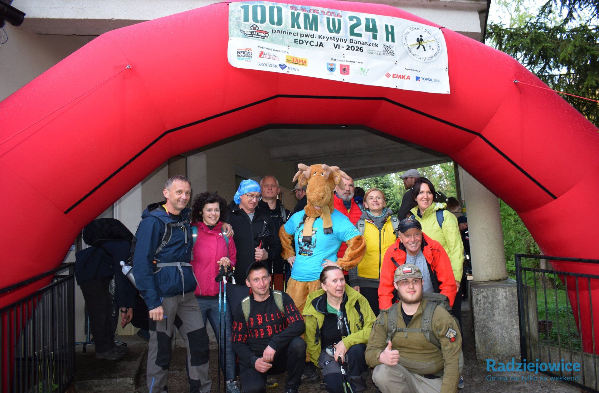 Grupa uczestników Marszu 100 km w 24 h pozuje do zdjęcia pod czerwonym łukiem startowym z banerem wydarzenia