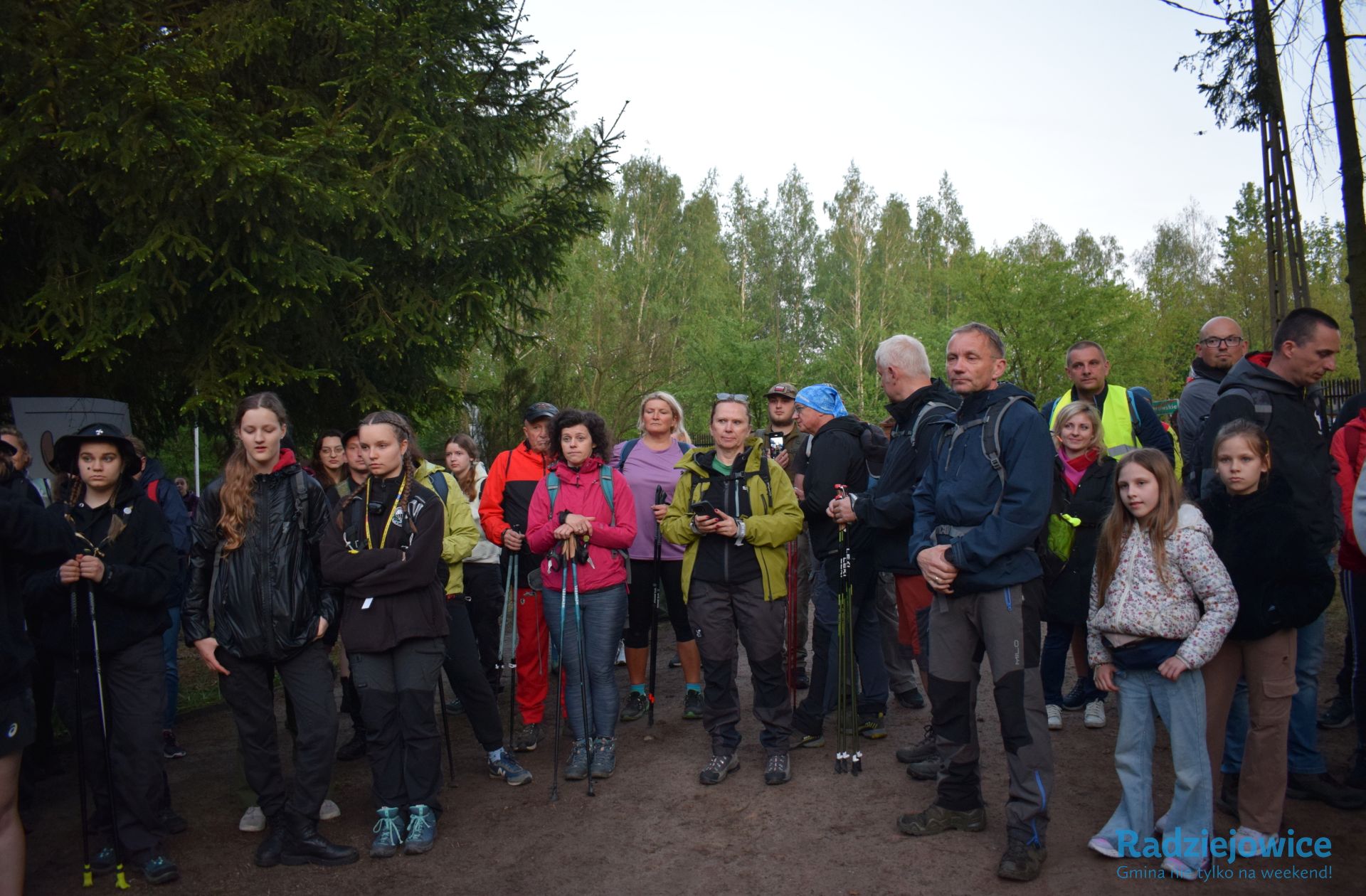 Grupa uczestników Marszu 100 km w 24 h zgromadzona przed startem trasy. Wśród uczestników znajdują się osoby dorosłe, dzieci i harcerze, część używa kijków trekkingowych