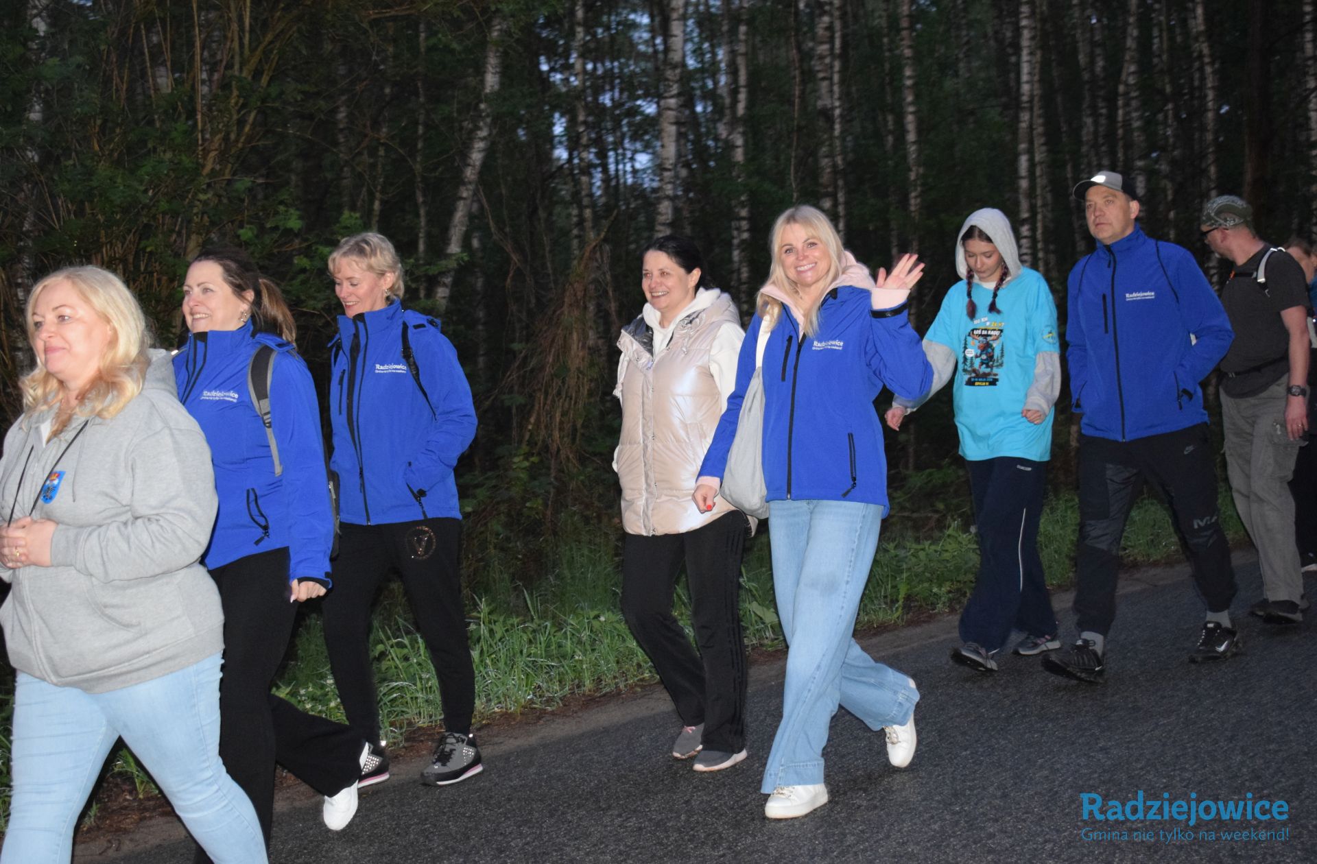 Grupa uczestników, w tym przedstawiciele gminy w niebieskich kurtkach, maszeruje drogą leśną podczas wydarzenia