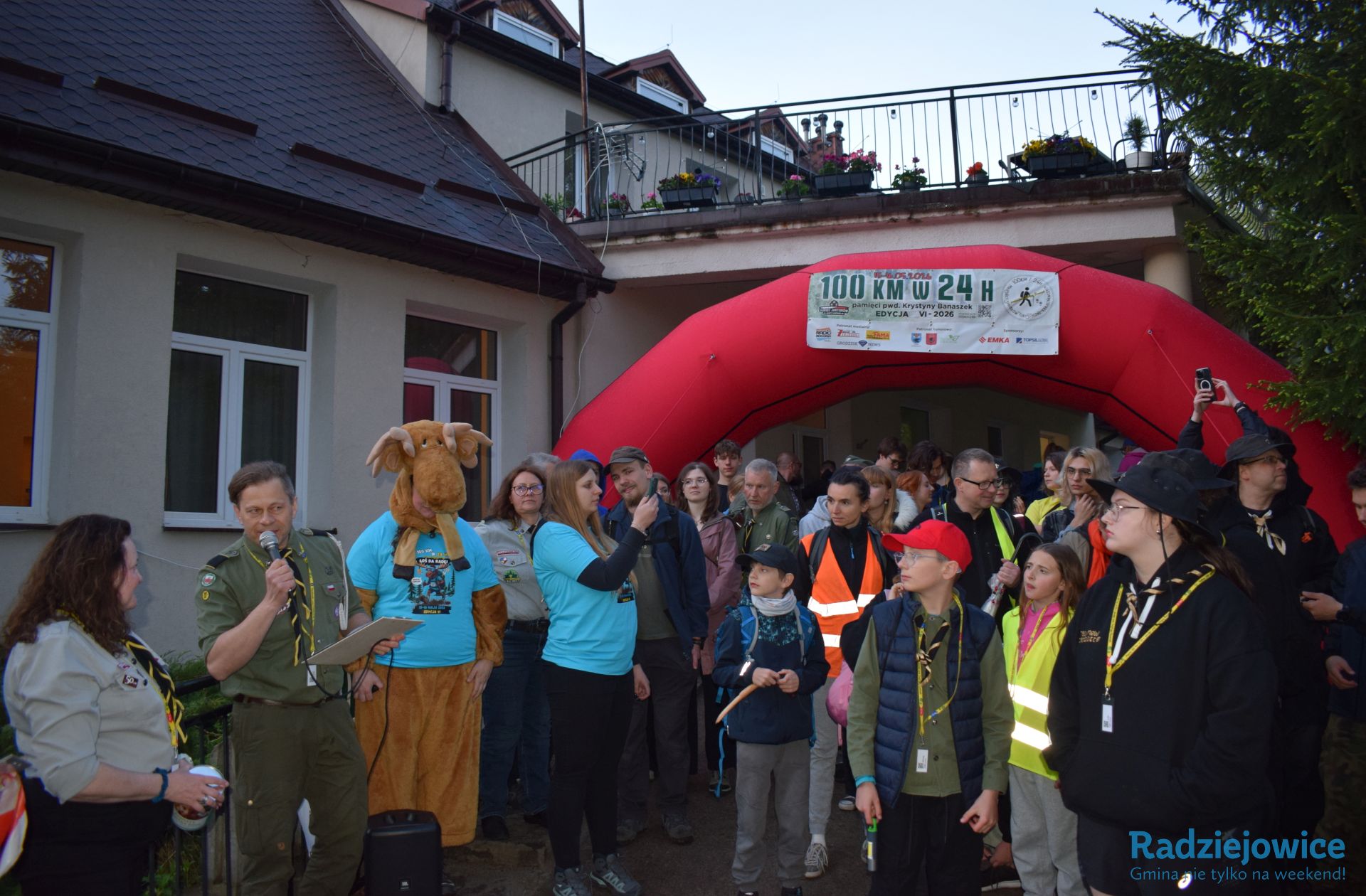 Organizator przemawia do uczestników Marszu 100 km w 24 h podczas rozpoczęcia wydarzenia. Obok stoją osoby w mundurach harcerskich oraz osoba w kostiumie łosia