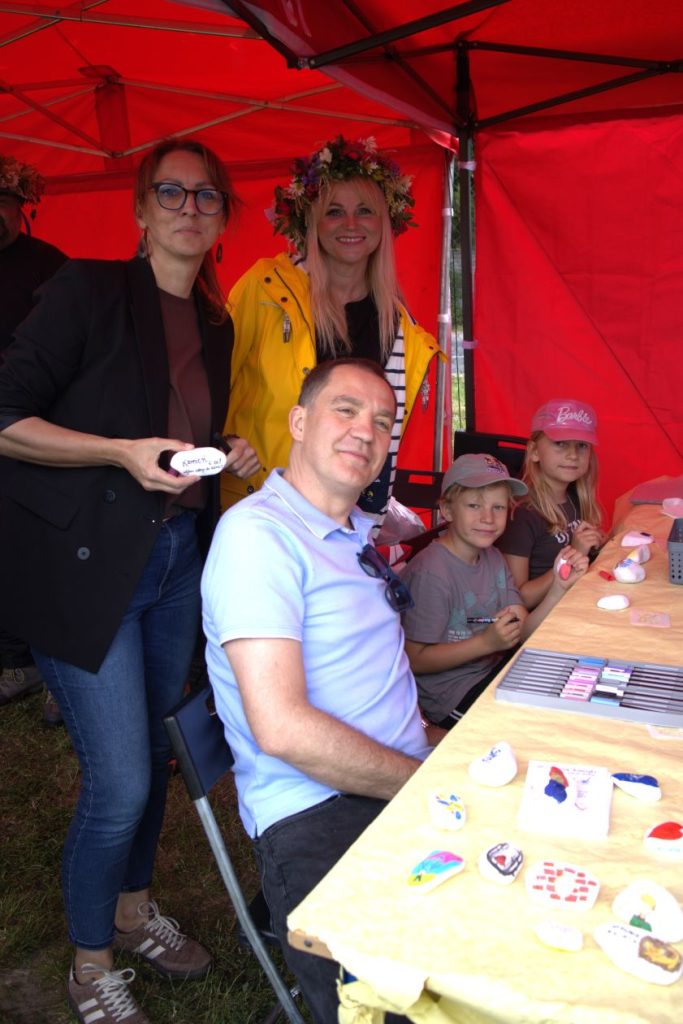 Dorośli i dzieci przy stole warsztatowym pod namiotem prezentują własnoręcznie ozdobione kamienie podczas rodzinnych zajęć plastycznych.