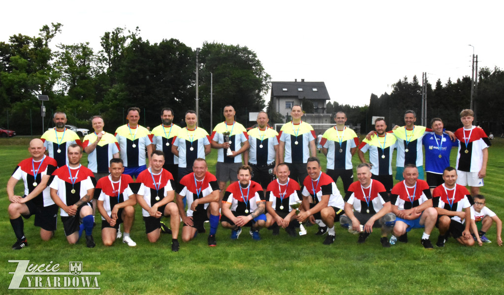 Drużyny sportowe pozują do wspólnego zdjęcia na boisku. Zawodnicy mają medale, a w centrum widoczne są puchary.