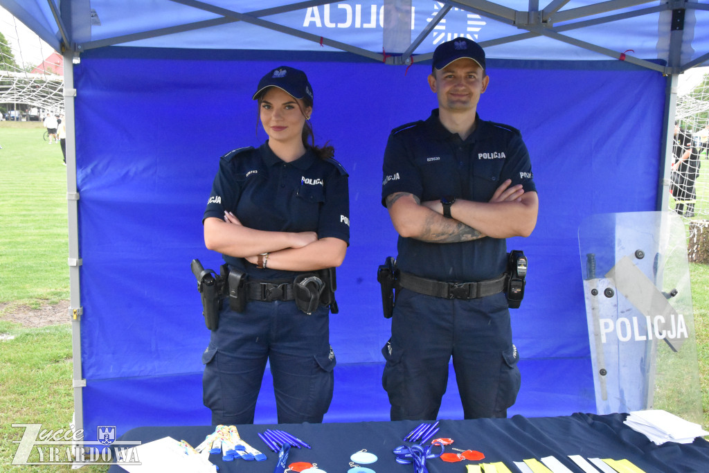 Dwoje funkcjonariuszy policji stoi przy stoisku promocyjnym pod namiotem. Na stole znajdują się materiały informacyjne i gadżety.