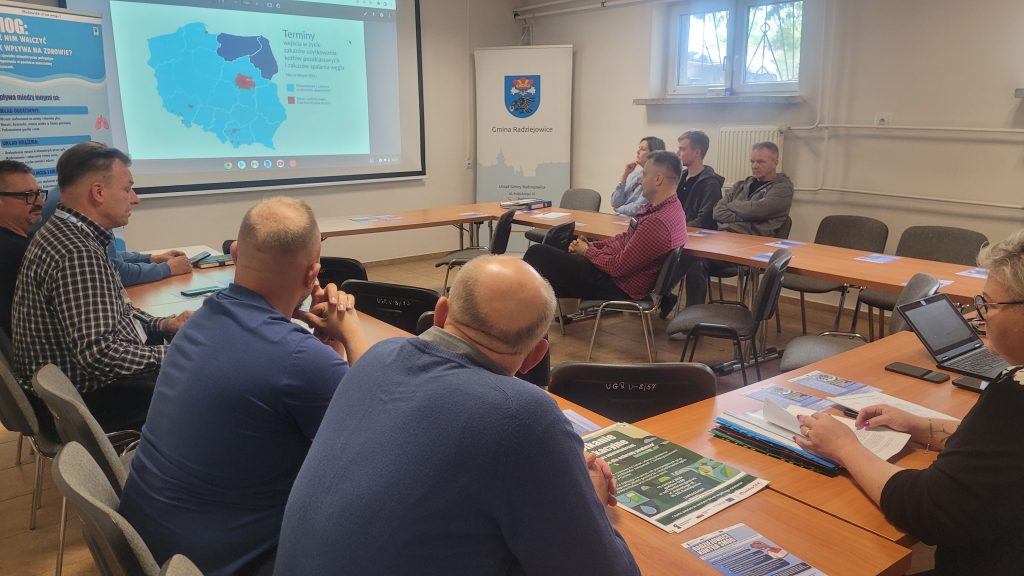 Uczestnicy spotkania siedzą przy stołach i uczestniczą w prezentacji dotyczącej ochrony powietrza. Na ekranie wyświetlona jest mapa Polski przedstawiająca terminy wejścia w życie zakazu użytkowania kotłów pozaklasowych.