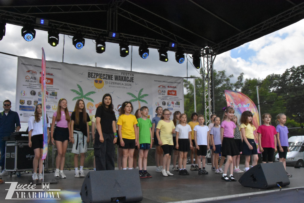 Grupa dzieci występuje na scenie. Uczestnicy ubrani są w kolorowe koszulki i stoją w rzędach.