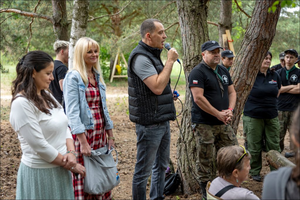 Grupa osób zgromadzona w lesie podczas wydarzenia plenerowego. Wójt trzyma mikrofon i przemawia do uczestników.