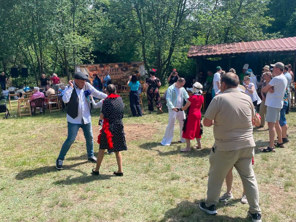 Kilka par tańczy na trawniku podczas wydarzenia plenerowego. W tle widoczna jest scena i uczestnicy spotkania.