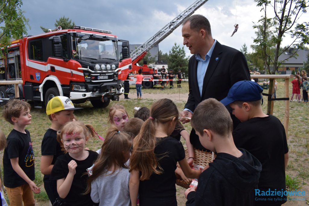 Przed wozem strażackim wojt gminy wręcza dzieciom słodkie upominki podczas obchodów dnia dziecka w szkole. W tle widoczne są pojazdy i sprzęt strażacki.