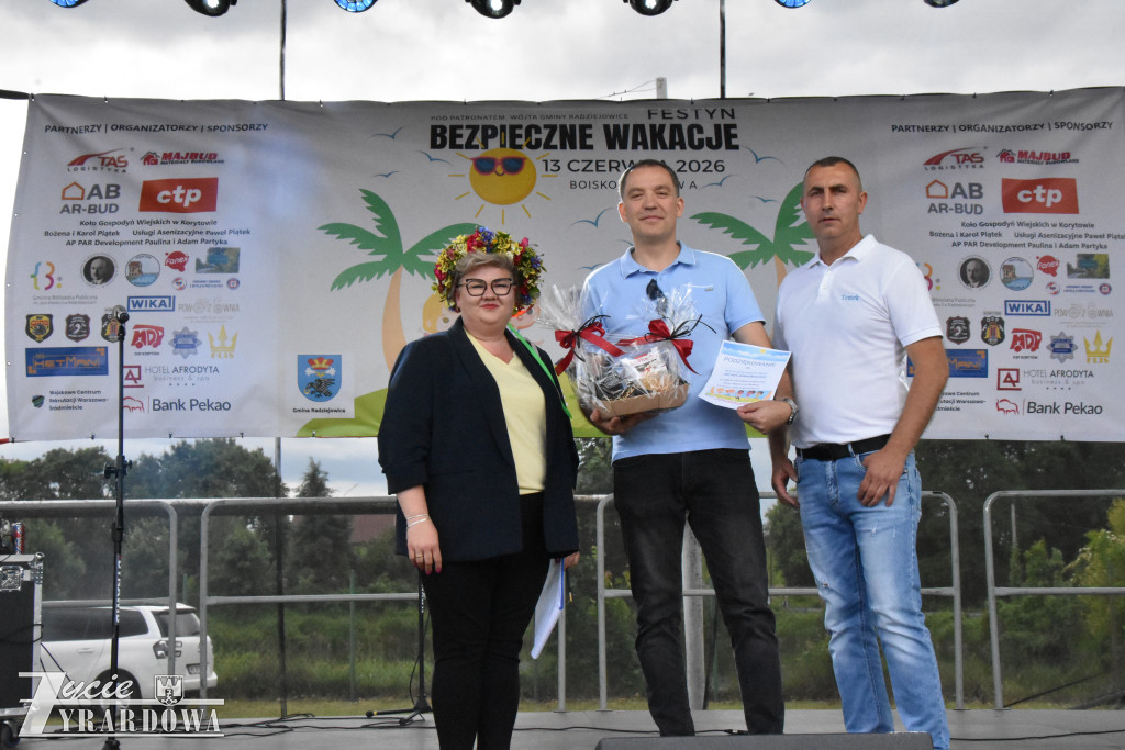 Trzy osoby pozują na scenie podczas wręczenia nagrody. Jedna z nich trzyma kosz upominkowy i dyplom.