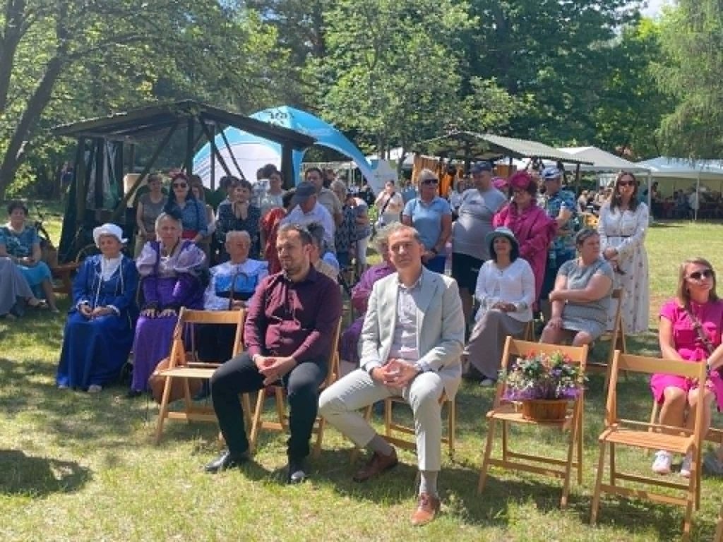 Uczestnicy siedzą na drewnianych krzesłach i stoją w tle podczas uroczystości plenerowej w otoczeniu drzew.