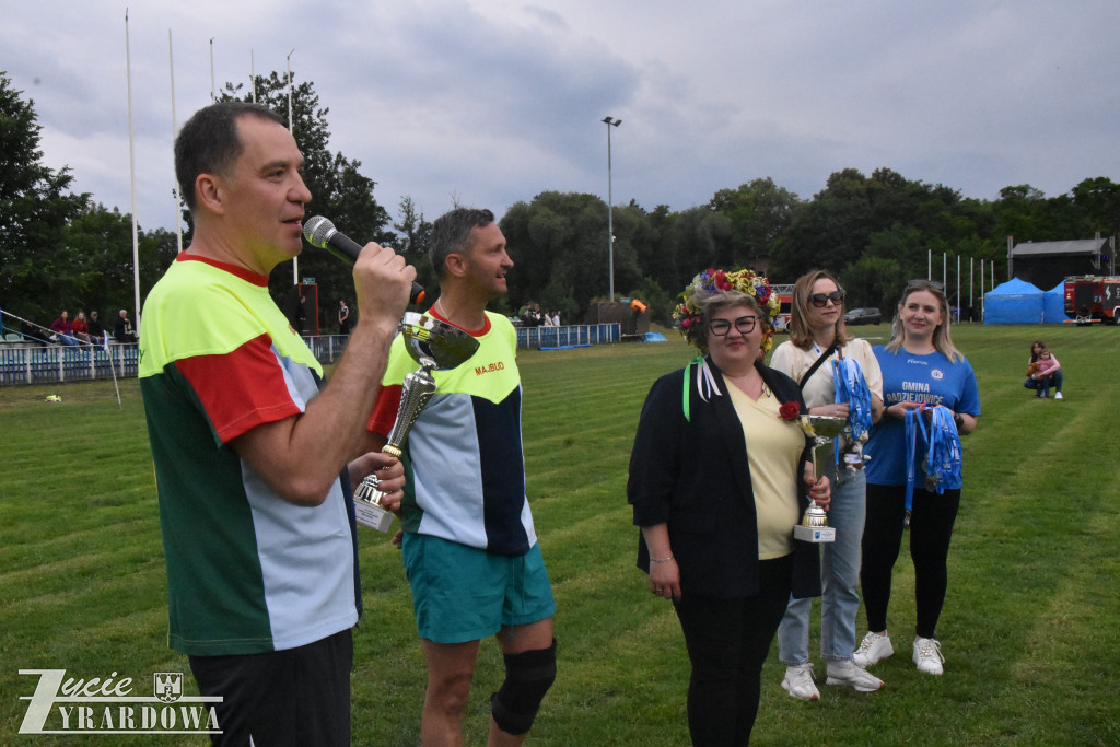 Uczestnicy wydarzenia stoją na boisku podczas ceremonii wręczenia nagród. Widoczne są puchary oraz osoby odbierające wyróżnienia.