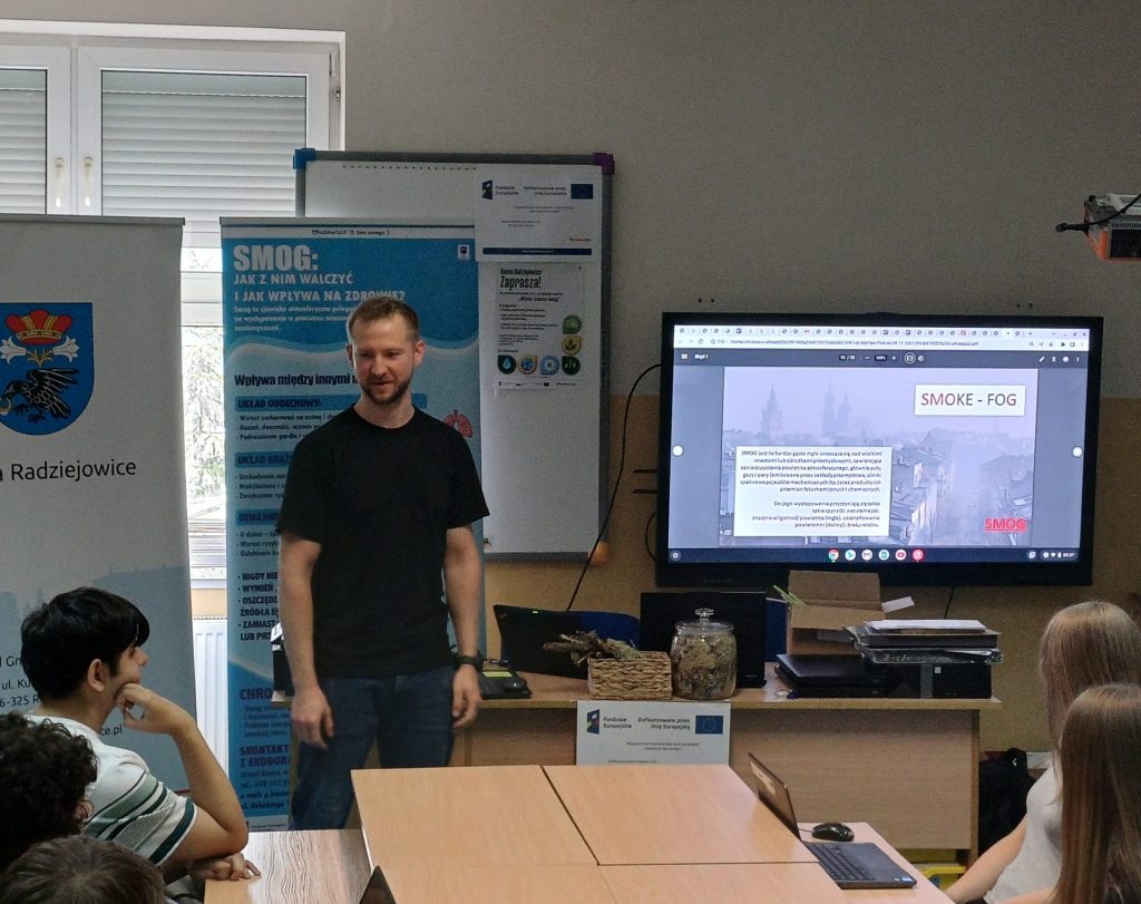 Prowadzący stoi przed grupą uczniów w sali lekcyjnej podczas spotkania edukacyjnego poświęconego jakości powietrza. Obok znajduje się roll-up informacyjny dotyczący smogu, a na monitorze wyświetlona jest prezentacja o zanie