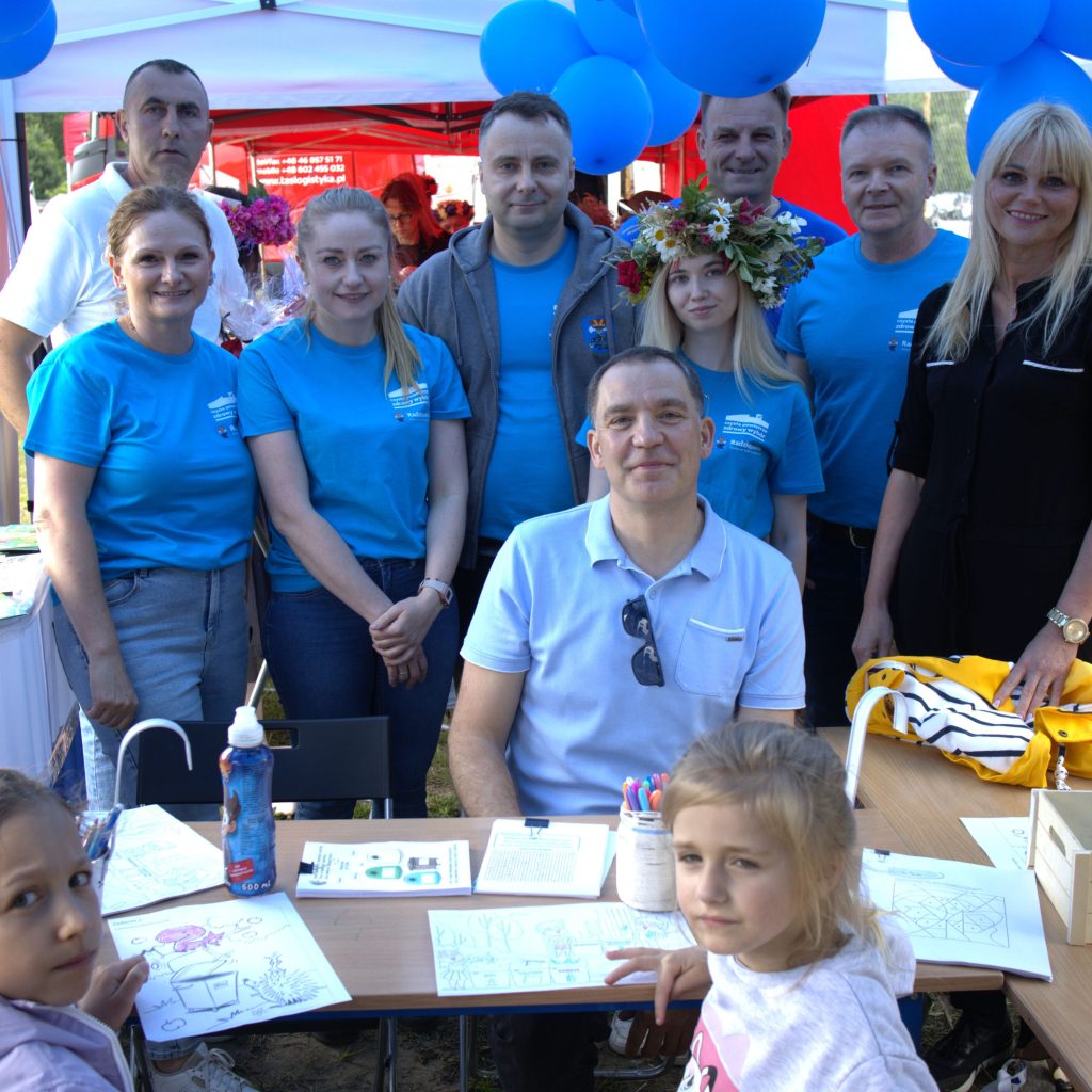 Grupa pracowników Urzędu w niebieskich koszulkach pozuje do zdjęcia przy stoisku informacyjnym. Na pierwszym planie dzieci siedzą przy stole z kolorowankami i materiałami do rysowania