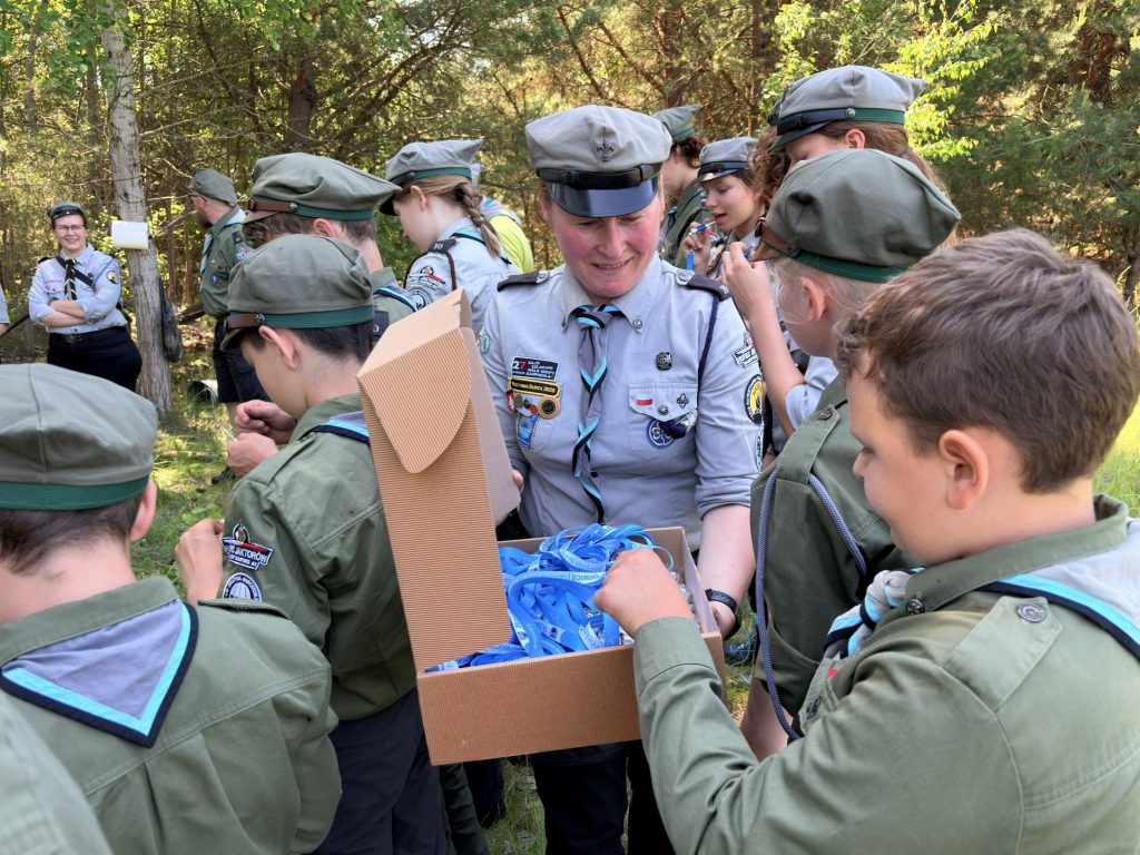 Instruktorka rozdaje harcerzom niebieskie smycze wyjmowane z otwartego pudełka w lesie.