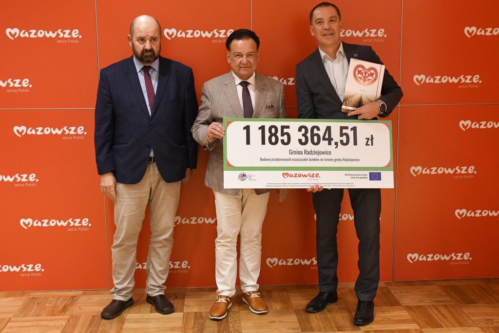 Na zdjęciu, wykonanym podczas oficjalnego wydarzenia, stoją trzy osoby na tle czerwonej ścianki z logo „Mazowsze serce Polski”. Po lewej stronie znajduje się Radosław Rybicki, Dyrektor Departamentu Rolnictwa i Rozwoju Obszarów Wiejskich. W centrum stoi Adam Struzik, Marszałek Województwa Mazowieckiego. Po prawej stronie znajduje się Wójt Gminy Radziejowice. Adam Struzik i Wójt trzymają dużą tablicę informacyjną przedstawiającą dofinansowanie dla Gminy Radziejowice w wysokości 1 185 364,51 zł na budowę przydomowych oczyszczalni ścieków. Wójt dodatkowo trzyma teczkę z okolicznościową grafiką.