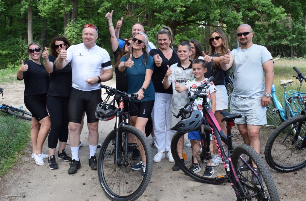 Grupa dorosłych i dzieci pozuje do wspólnego zdjęcia z rowerami na leśnej ścieżce. Kilka osób pokazuje gest uniesionego kciuka.