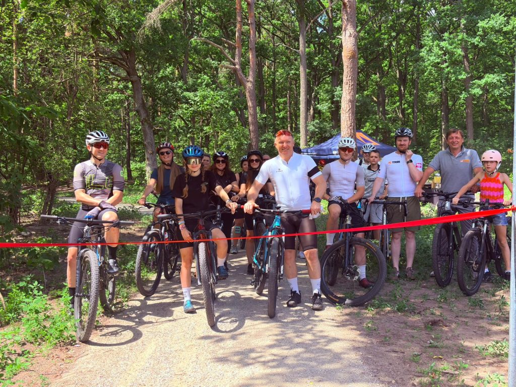 Grupa uczestników na rowerach stoi przed czerwoną wstęgą na leśnej ścieżce rowerowej. W tle znajdują się drzewa oraz namiot organizatorów.