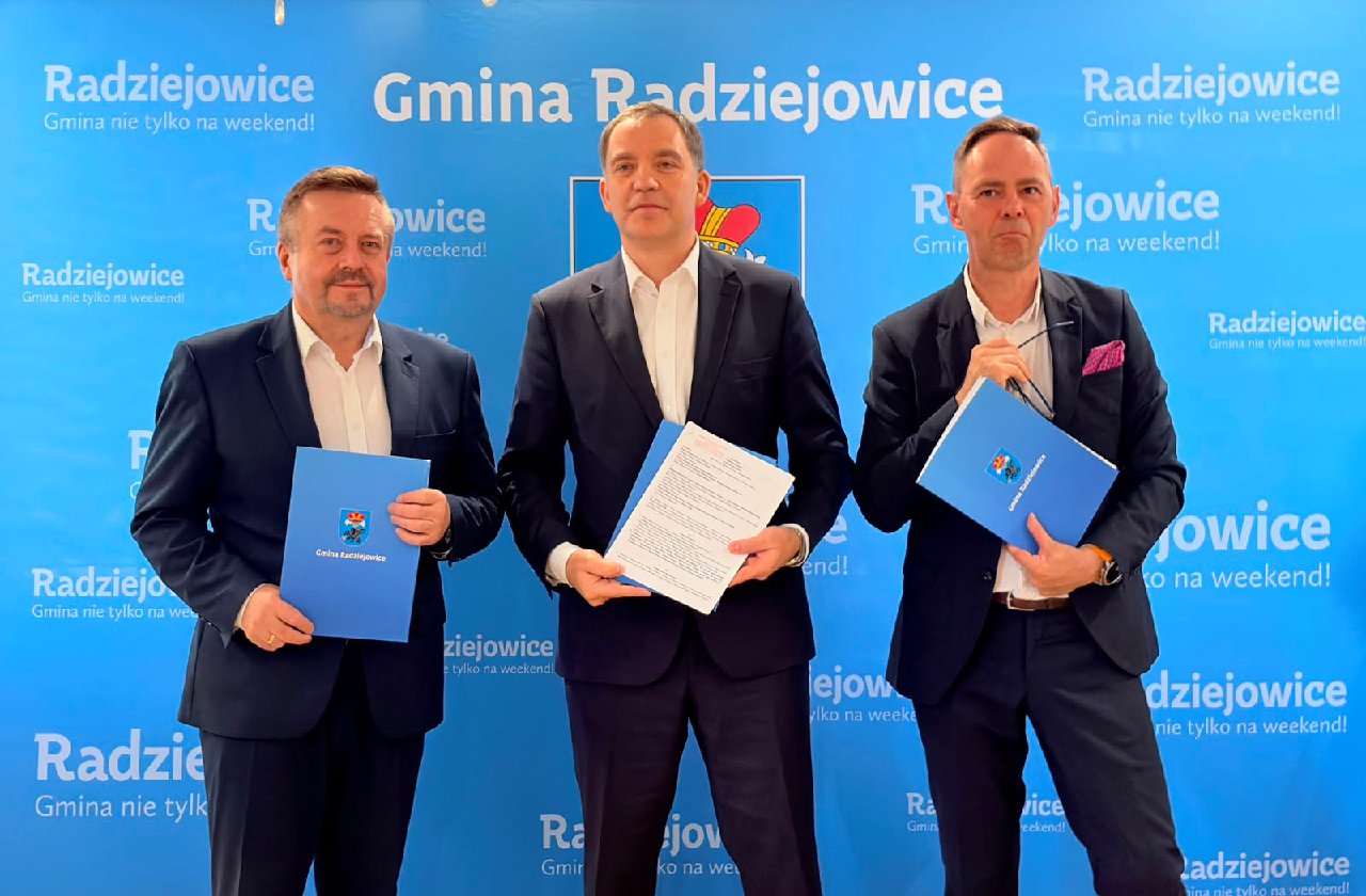 trzech Panów pozuje do zdjęcia tzrymając niebieskie teczki z dokumentami. w tle niebieska ścianka z herbem Gminy Radziejowice i białymi napisami "Gmina Radziejowice"