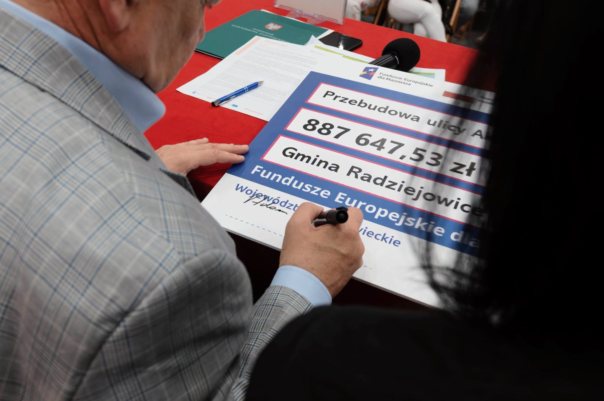 Osoba podpisuje tablicę informującą o dofinansowaniu przebudowy ulicy Akacjowej w Gminie Radziejowice z programu Fundusze Europejskie dla Mazowsza 2021–2027. Na stole znajdują się dokumenty związane z podpisaniem umowy.