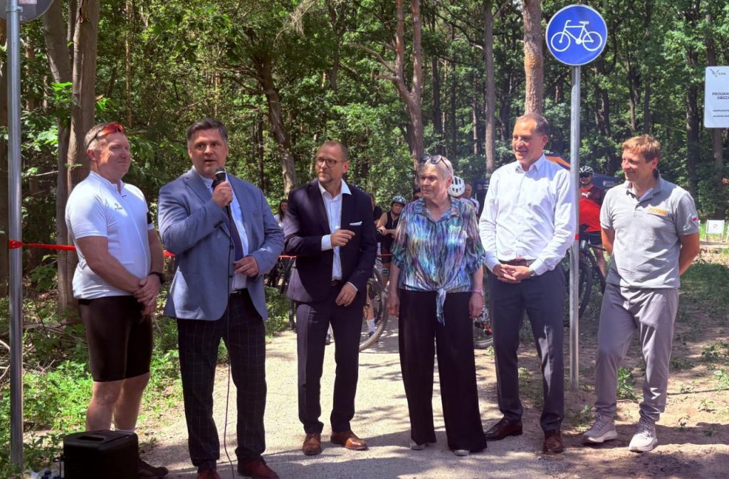 Przedstawiciele organizatorów i zaproszeni goście stoją na leśnej ścieżce podczas przemówienia. W tle widoczny jest znak drogi dla rowerów oraz uczestnicy wydarzenia.