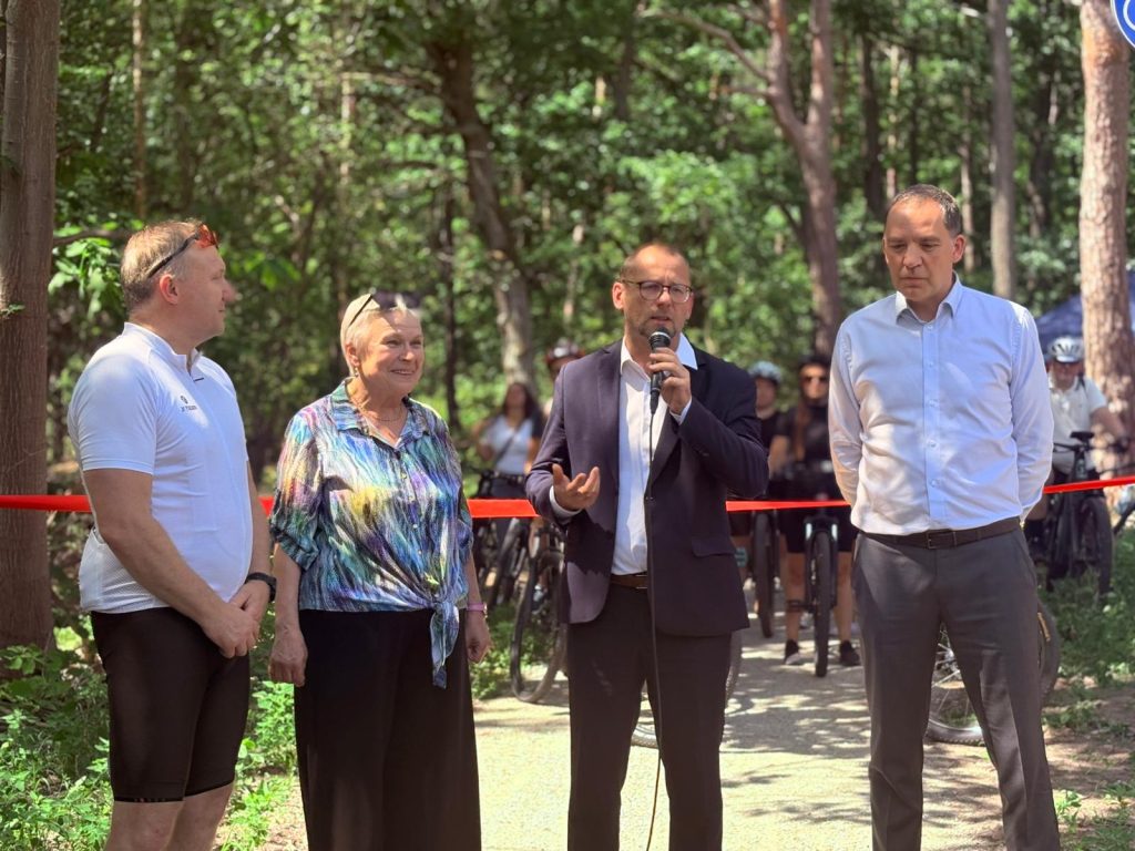 Przemawiający trzyma mikrofon podczas uroczystości otwarcia leśnej ścieżki rowerowej. Obok stoją pozostali uczestnicy wydarzenia.
