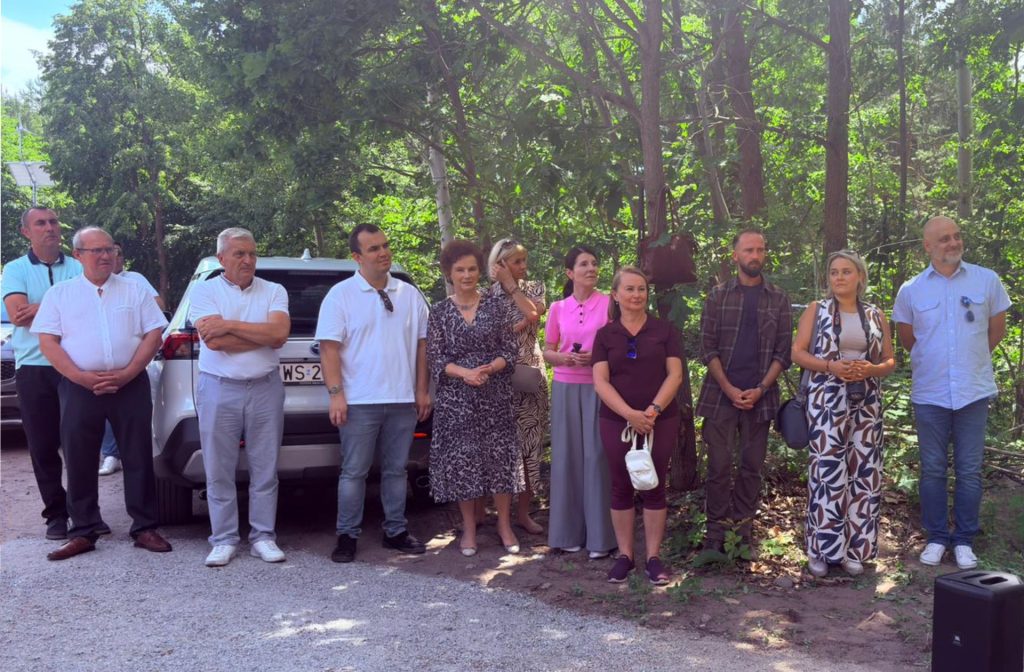 Uczestnicy uroczystości stoją wzdłuż leśnej drogi i przysłuchują się przemówieniom. W tle widoczne są zaparkowane samochody.