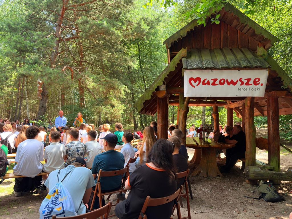 Uczestnicy wydarzenia „Świadkowie Historii” słuchają prelekcji na terenie skansenu. W tle widoczna jest wiata z banerem „Mazowsze. Serce Polski”.