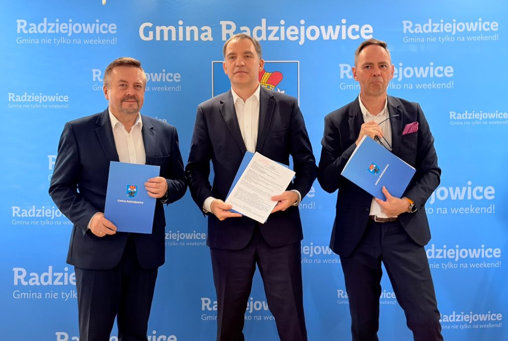 trzech Panów pozuje do zdjęcia tzrymając niebieskie teczki z dokumentami. w tle niebieska ścianka z herbem Gminy Radziejowice i białymi napisami "Gmina Radziejowice"