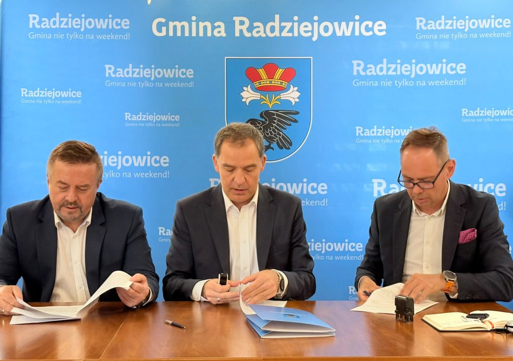 trzy osoby siedzą przy biurku i podpisują dokumenty. w tle niebieska ścianka z herbem Gminy Radziejowice i białymi napisami "Gmina Radziejowice"