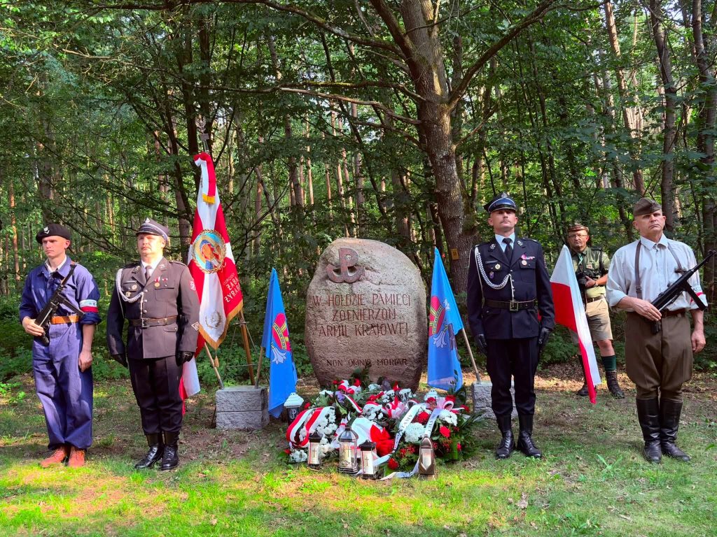 Przed pomnikiem poświęconym żołnierzom Armii Krajowej w lesie stoi honorowa warta w historycznych i strażackich mundurach. U stóp pomnika znajdują się biało-czerwone wiązanki, znicze oraz flagi Polski i Gminy Radziejowice.