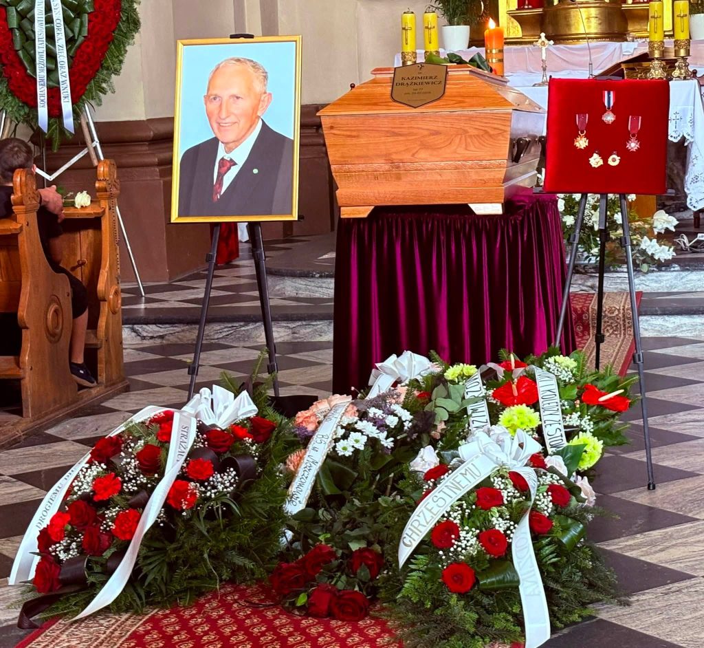 Wnętrze kościoła. Na środku znajduje się trumna śp. Kazimierza Drążkiewicza ustawiona przed ołtarzem. Obok stoi portret zmarłego na sztaludze oraz tablica z odznaczeniami. Przed trumną ułożone są liczne wieńce i wiązanki kwiatów.