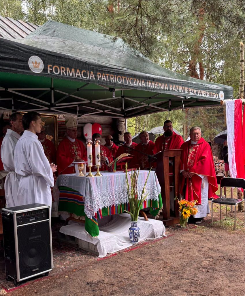 Msza polowa odprawiana przy drewnianej kaplicy w lesie. Przy ołtarzu znajduje się kilku duchownych, a przed nim ministranci. Nad ołtarzem widoczny napis „Formacja Patriotyczna imienia Kazimierza Wielkiego”.
