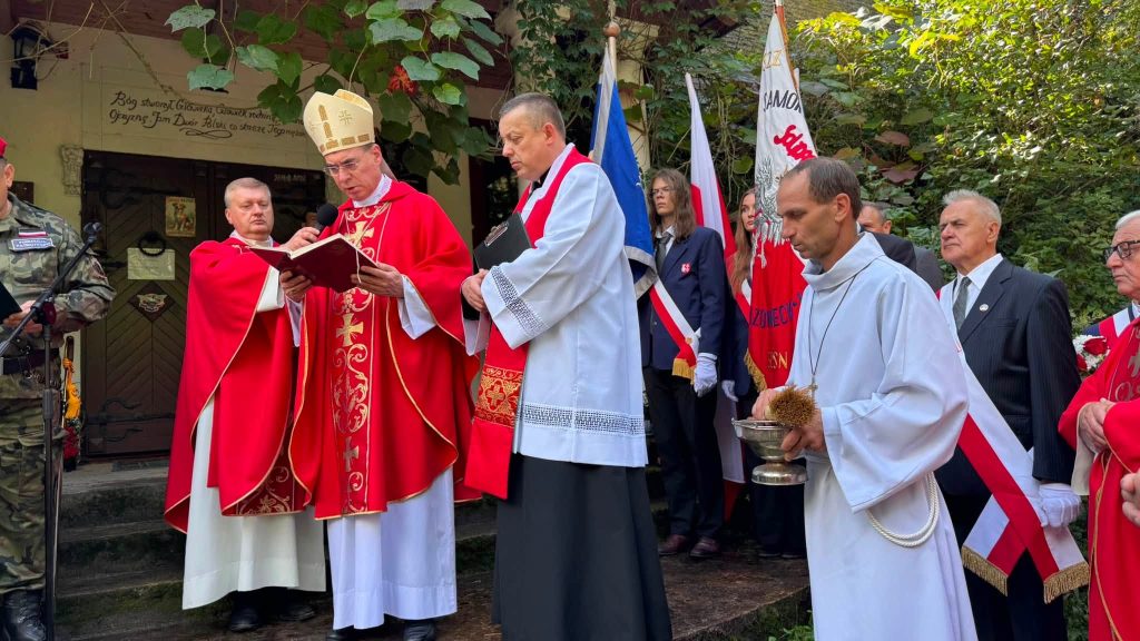 Duchowny w czerwonych szatach liturgicznych odprawia nabożeństwo przed wejściem do drewnianej kaplicy. W tle widoczne są poczty sztandarowe.