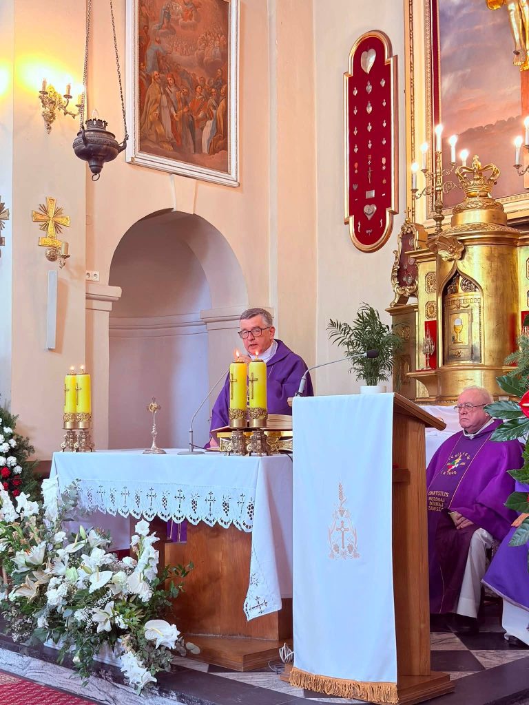 Kapłan przewodniczący uroczystości wygłasza homilię przy ołtarzu. W prezbiterium widoczne są świece, kwiaty oraz drugi duchowny siedzący obok.