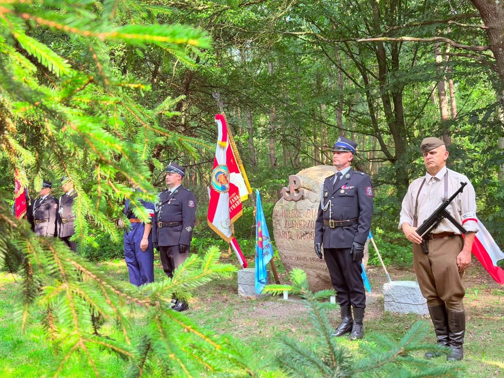 Honorowa warta stoi przed pomnikiem żołnierzy Armii Krajowej. Zdjęcie wykonano zza gałęzi świerku, które częściowo zasłaniają pierwszy plan.