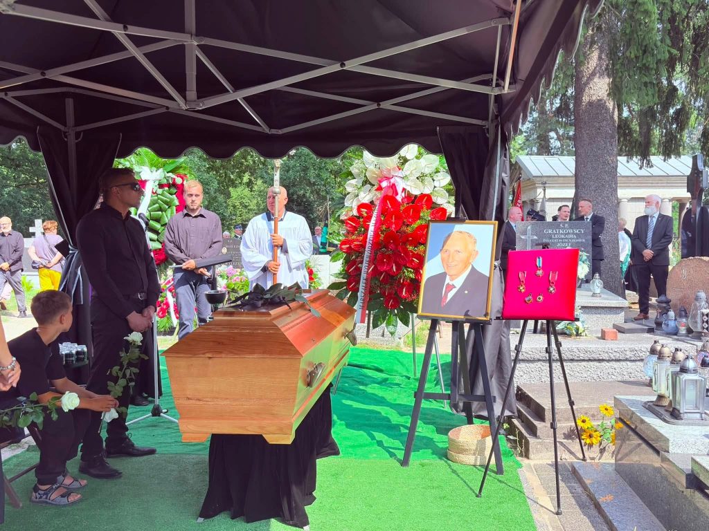 Na cmentarzu trumna śp. Kazimierza Drążkiewicza znajduje się pod namiotem przygotowanym do ceremonii pochówku. Obok ustawiono portret zmarłego, tablicę z odznaczeniami oraz wieńce pogrzebowe. W tle stoją uczestnicy uroczystości.
