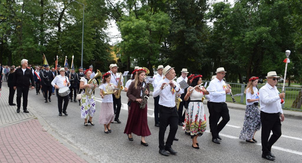 Uroczysty przemarsz ulicami Radziejowic. Orkiestra dęta w białych koszulach i kapeluszach idzie na czele, a za nią podążają przedstawiciele służb i uczestnicy wydarzenia.
