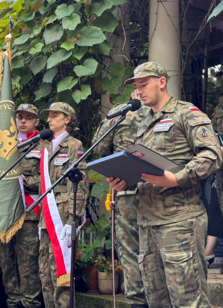 Umundurowany żołnierz przemawia do uczestników uroczystości przy mikrofonie. W tle stoją członkowie pocztu sztandarowego.