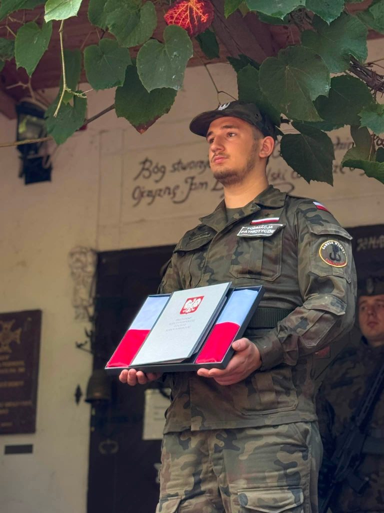 Umundurowany przedstawiciel formacji patriotycznej prezentuje pamiątkowe wyróżnienie w biało-czerwonych barwach.