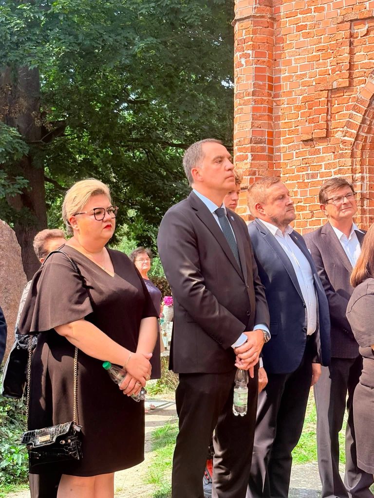 Na cmentarzu podczas ceremonii pogrzebowej stoją przedstawiciele władz samorządowych oraz uczestnicy uroczystości. W tle widoczny jest ceglany mur kościoła i drzewa.