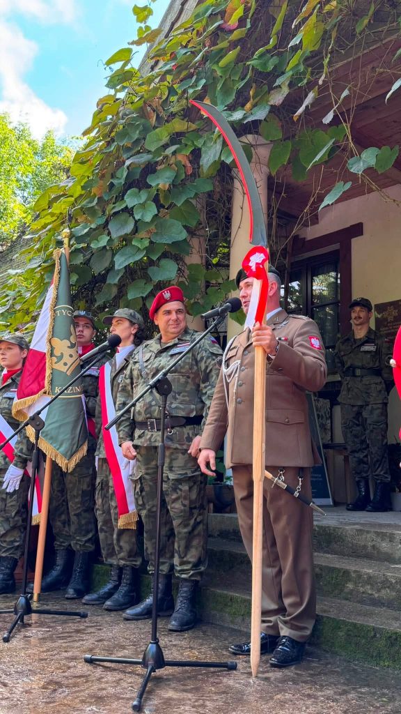 Umundurowany przedstawiciel formacji patriotycznej przemawia przy mikrofonie, trzymając historyczną broń. Za nim stoją członkowie pocztów sztandarowych.