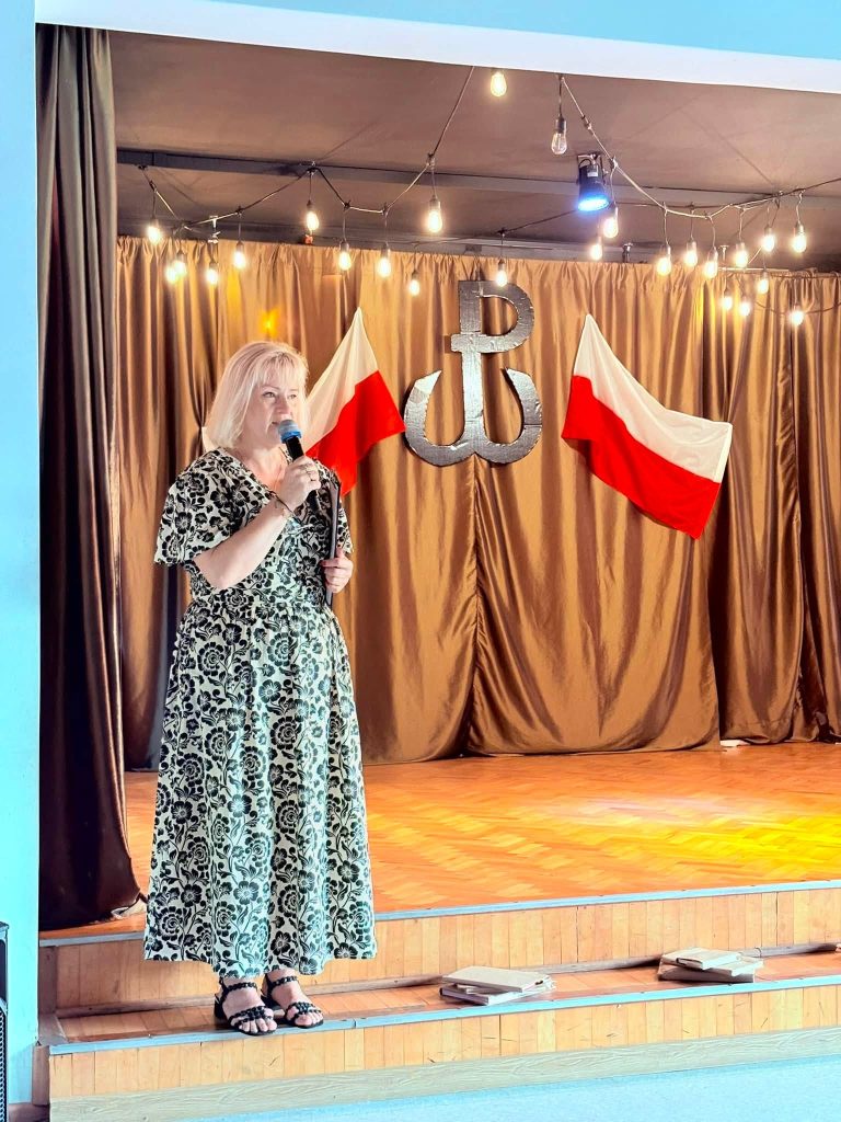 Kobieta przemawia na scenie podczas części artystycznej obchodów. W tle znajdują się dwie flagi Polski oraz symbol Polski Walczącej.