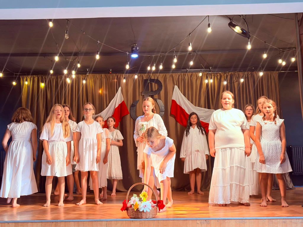 Grupa dzieci w białych strojach prezentuje występ artystyczny na scenie podczas obchodów rocznicowych.