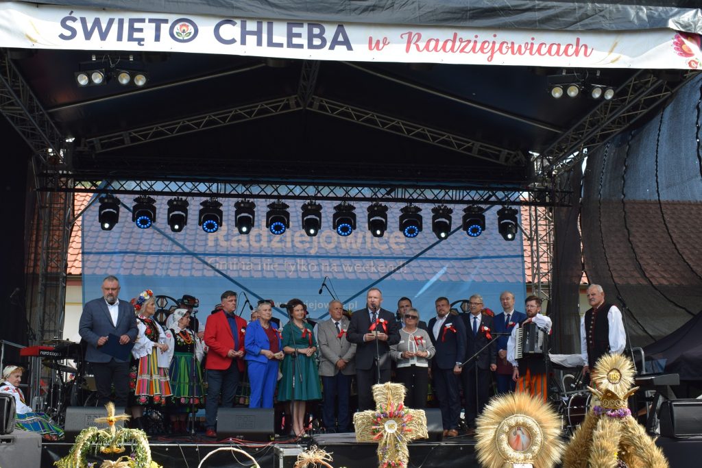 Uczestnicy Święta Chleba stoją na scenie pod banerem z nazwą wydarzenia. Na pierwszym planie znajdują się tradycyjne wieńce dożynkowe, a na scenie przedstawiciele organizatorów i lokalnych społeczności.